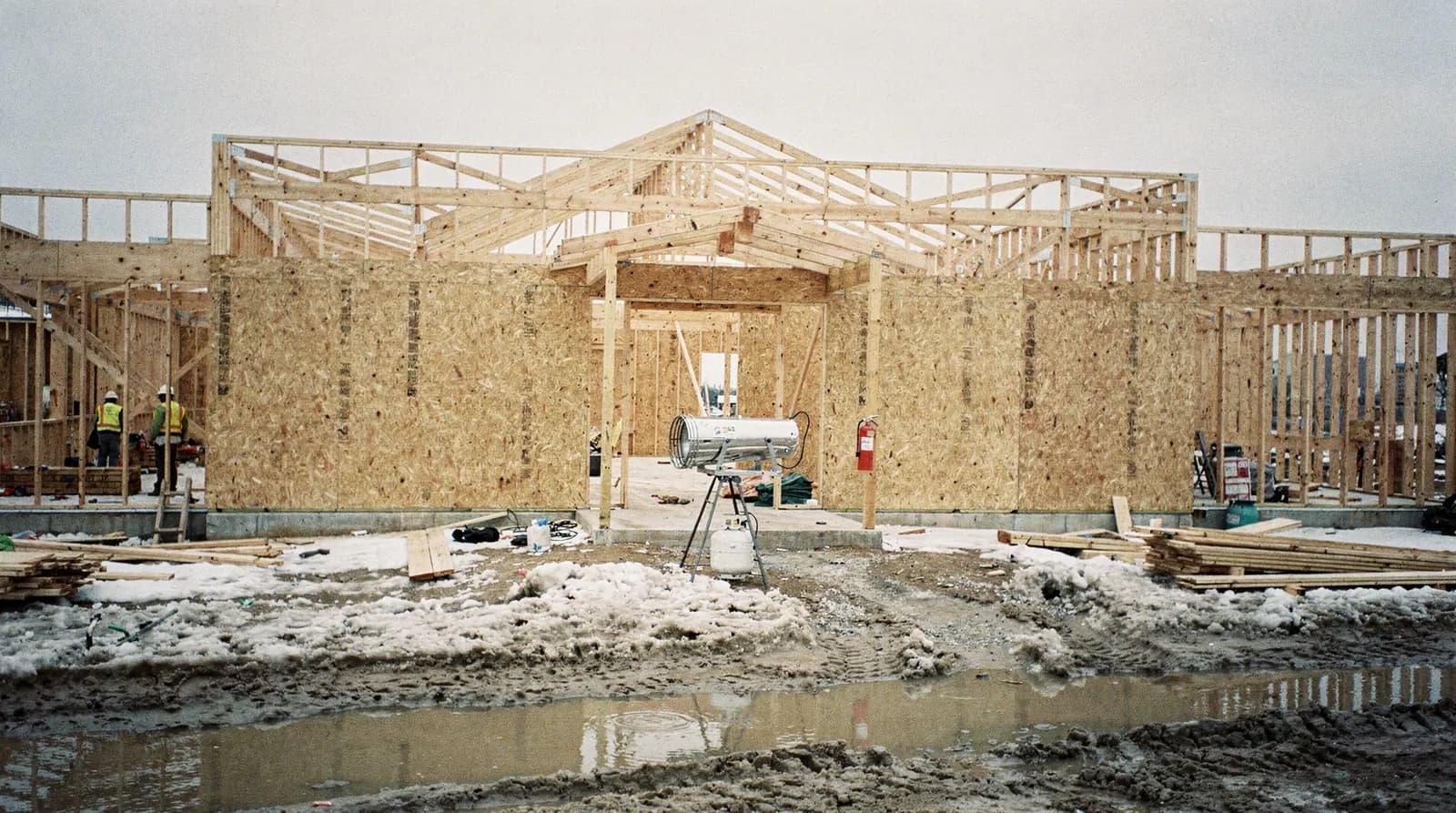 A partly framed commercial building on a winter afternoon with temporary site fencing, a laydown area and a fire extinguisher station beside the site gate