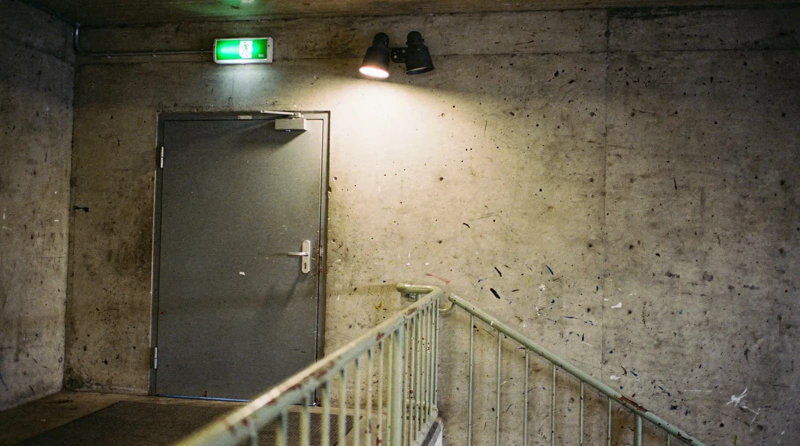 A facility worker on a step ladder checking a battery emergency light unit above a corridor door beside an exit sign
