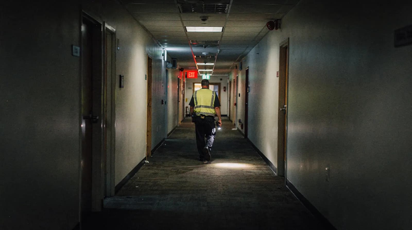 A person in a hi-vis vest walking a quiet building corridor with a flashlight at night, past a fire extinguisher cabinet and an exit sign