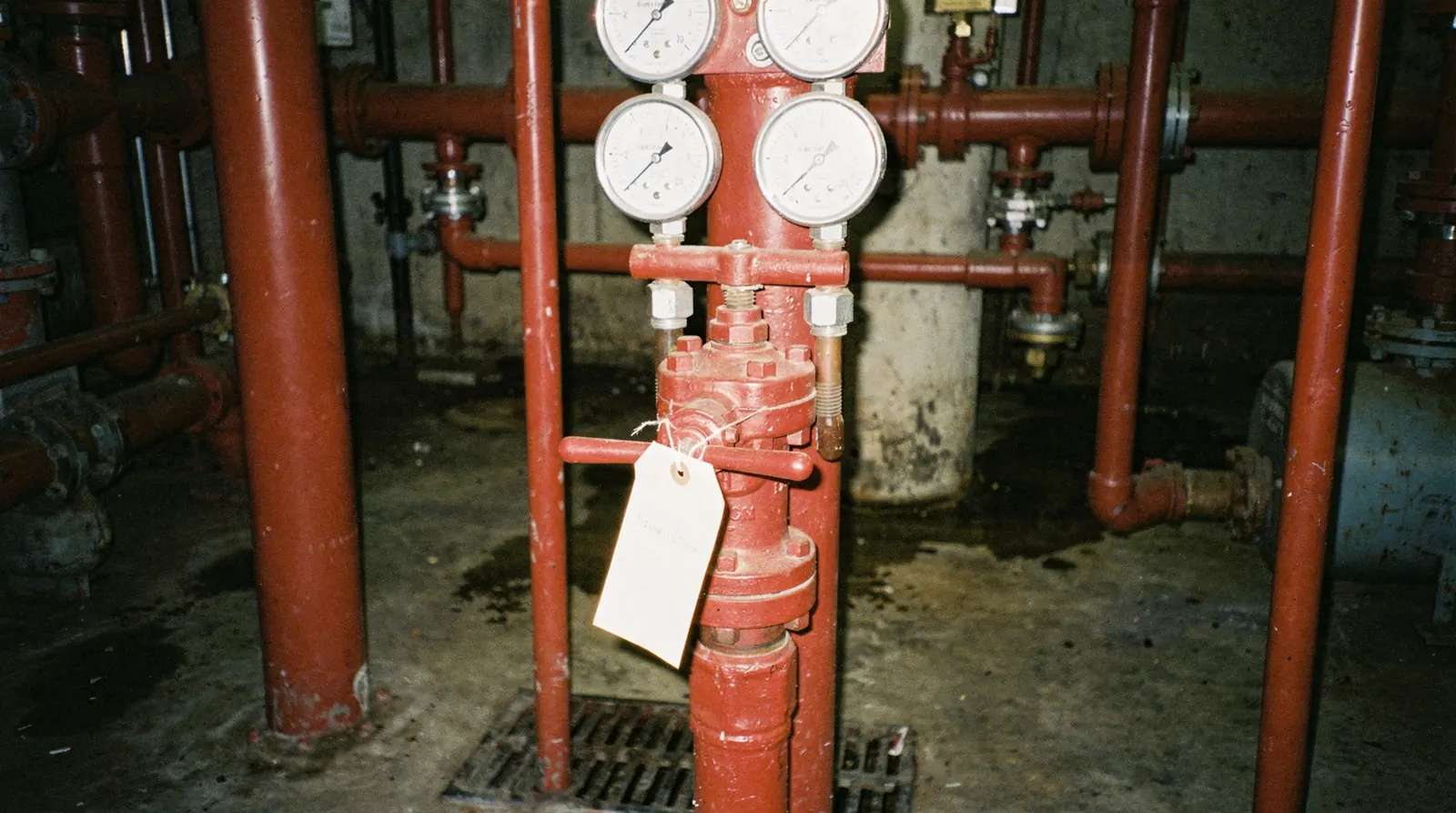 A closed and tagged sprinkler control valve on a riser in a plain utility room, with a clipboard hanging beside it